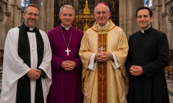 Four Anglican and Catholic clergy members standing together inside a cathedral, wearing traditional religious robes, with a crucifix and stained-glass windows behind them.